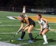 Two female field hockey players compete for the ball on a brightly lit turf field, showcasing athletic skill and teamwork.