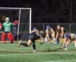 Field hockey players compete under stadium lights, with a goalie preparing to defend against a shot on goal.