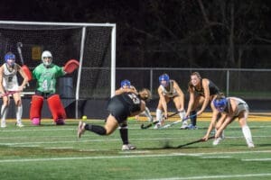 Field hockey players compete under stadium lights, with a goalie preparing to defend against a shot on goal.