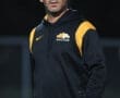 Soccer coach in a black and yellow tracksuit, focused and walking on the field during practice.