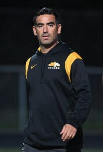 Soccer coach in a black and yellow tracksuit, focused and walking on the field during practice.