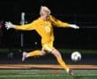 Goalkeeper in a yellow jersey diving to kick a soccer ball during an intense night game on the field.