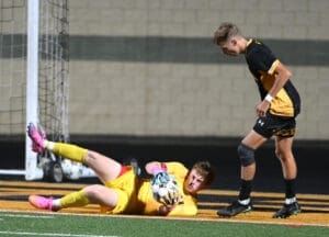 A goalkeeper makes a save while a teammate watches during a night soccer match on a turf field.