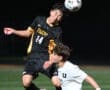 High school soccer players contest a header during a night game on the field. The scoreboard is visible in the background.