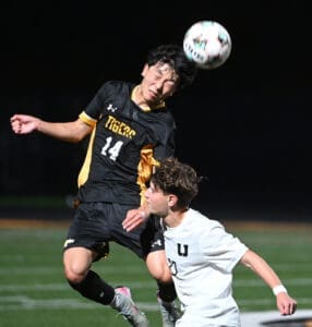 High school soccer players contest a header during a night game on the field. The scoreboard is visible in the background.