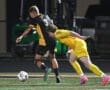High school soccer match with a player in black dribbling past a yellow-clad goalkeeper attempting to block the ball.
