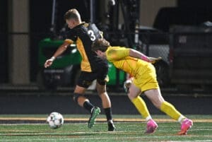 High school soccer match with a player in black dribbling past a yellow-clad goalkeeper attempting to block the ball.