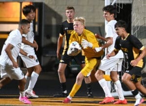 High school soccer match with players clustered around the goal, goalkeeper in yellow preparing to catch the ball.
