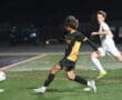 High school soccer match where a player in black and yellow kicks the ball, with an opponent in white nearby.