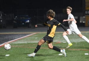 High school soccer match where a player in black and yellow kicks the ball, with an opponent in white nearby.